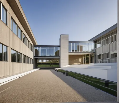 a building with straight lines, with colored panels on the facade, windows with dark frames, in the background a glass passage with a concrete pillar in the center and trees behind, a garden on the ri