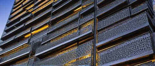 Modern building exterior, architectural wire mesh panels, silver metallic finish, intricate geometric patterns, industrial chic aesthetic, urban cityscape, skyscraper, evening time, warm golden lighti