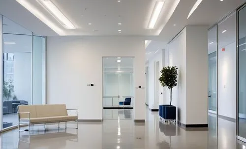 A hospital wing entrance with white walls and stone floors. There is a navy blue sofa. There are glass double doors in the center. There are ceiling lights in the hallway. The room has a  calm atmosph