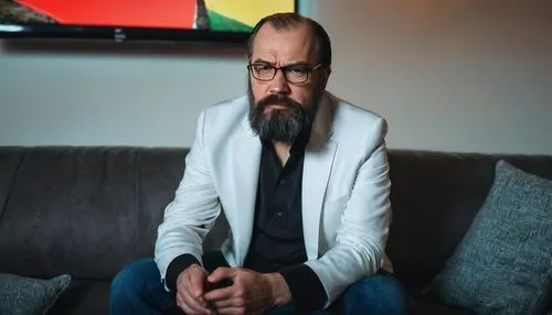 Netflix founder, Lenin-like beard, mature man, 50yo, serious facial expression, black hair, glasses, white shirt, black blazer, jeans, sitting on a couch, watching TV, remote control in hand, backgrou