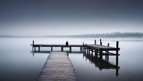 An old, weathered wooden pier extending out into a calm, misty lake. The horizon is barely visible, shrouded in a delicate fog that diffuses the soft morning light. Standing at the end of the pier is 