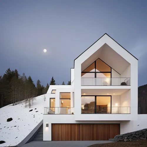 a house in the mountains, black zinc sloping roof, white plaster on the facade, with wooden slatted details, large windows, two balconies, garage below with two gates, outdoor patio on the left side w