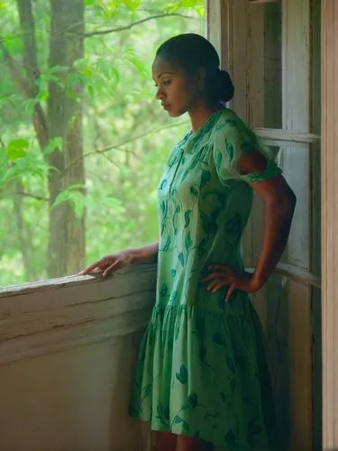 This picture shows a pretty Creole woman in a light-colored dress standing at a window. Her gaze is directed outside and she appears to be pensive. Bright colors. The surroundings outside appear green