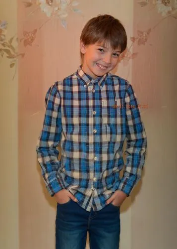 The image shows a young boy with short brown hair and brown eyes, smiling and looking at the camera. He is wearing a blue, white, and yellow plaid button-down shirt with a collar. He also has on a pai