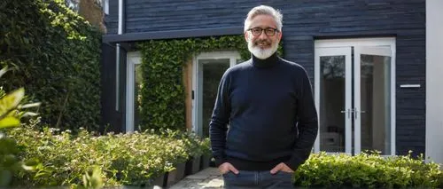 Middle-aged male architect, bespectacled, grey hair, trimmed beard, wearing a black turtleneck sweater, dark blue jeans, brown leather shoes, holding a large portfolio, standing in front of a modern T