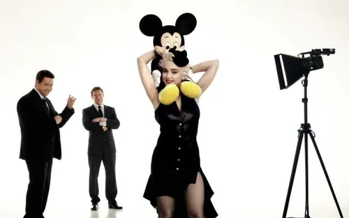 Madonna is on a movie set and she has Mickey Mouse on her shoulders. Madonna is wearing a sexy dress and has beautiful legs and she is wearing high heels. The movie set is full of crew members such as