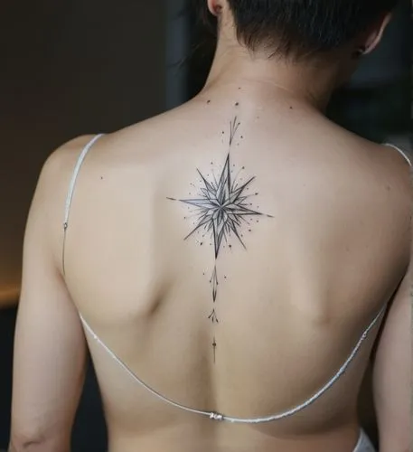 The image shows a man with a delicate geometric pattern tattooed on his back. The tattoo is located on the upper back, below the neck. The design is a stylized compass or star, with a center point and