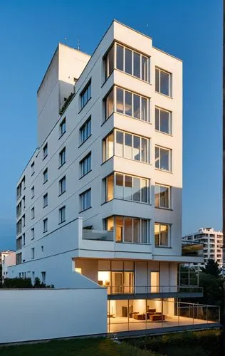 The image shows a tall residential building with a modern design and large windows. There are balconies on each floor, and the building has a white exterior with wood accents.,seidler,escala,plattenba