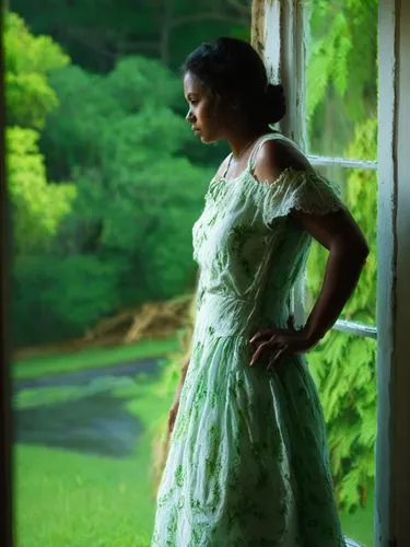 This picture shows a pretty Creole woman in a light-colored dress standing at a window. Her gaze is directed outside and she appears to be pensive. Bright colors. The surroundings outside appear green