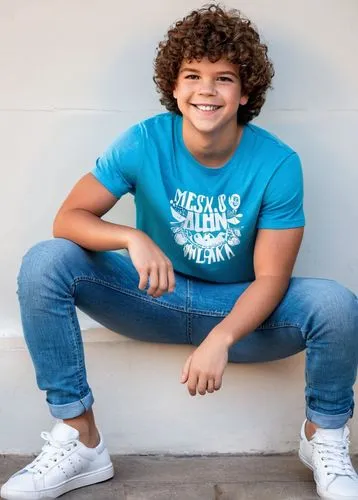 Gaten Matarazzo, young boy, smiling face, bright blue eyes, messy brown hair, casual wear, white graphic t-shirt, light blue jeans, sneakers, relaxed pose, leaning on a wall, indoor setting, warm ligh