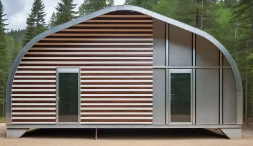 A quonset hut house, with wooden beams and a steel shell. The beams on the right are metal with glass paneling behind it and the doors are on either side. the house is located in a forest with a concr