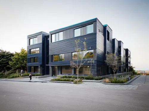 Modern building with two massing, bluish metal slatted facade on the right side, dark stone on the left side, floor to ceiling glass in-between the two , small punch windows on the front side of the b