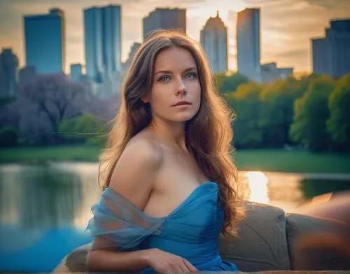 a young caucasian woman  brown long hair blue eyes wearing a blue dress laying in a sofa in central park whit a lake and building at sunset as background ,romantic portrait,girl on the river,portrait 