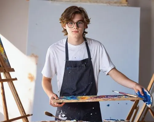 young artist boy, 18yo, messy brown hair, bright blue eyes, glasses, white shirt with paint stains, black apron, jeans, Converse shoes, holding a palette, standing in front of an easel, studio backgro