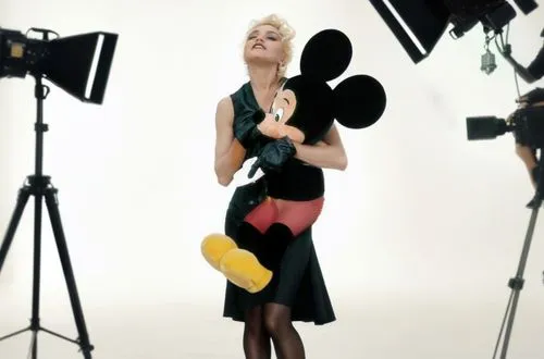 Madonna is on a movie set with Mickey Mouse in her arms! But Donna has short, blonde, platinum hair. Madonna is wearing a sexy dress and has beautiful legs. The movie set is full of crew members such 