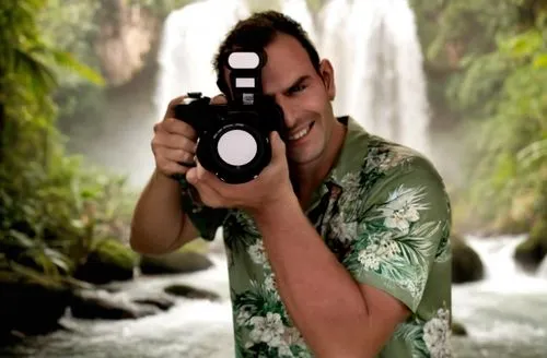 the photographer raises his arm while in the background there is a jungle with a waterfall. his mouth is closed but he is smiling.,a man with a camera taking a po in front of a waterfall,nature photog