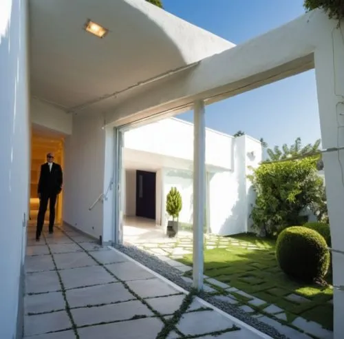 INTERIOR DEL VESTIBULO DE UNA CASA, QUE DA  AUN JARDIN INTERIOR CON MACETAS Y PLANTAS EXOTICAS, CANCALES DE ALUMINIO, CRISTAL, ILUMINACIÓN ELEGANTE Y TENUE,a man is walking in an open doorway into a b