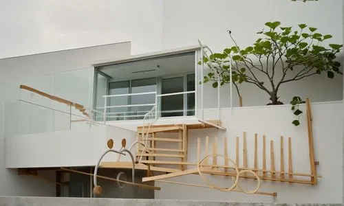 UNA CASA  ESTILO MINIMALISTA CON UN MURO DECORADO CON UNA JARDIENRA CON PLANTAS DE FORMIO, UN GRAN ARBOL SALE DE LA BARDE QUE LIMITA LA CASA, UNA GRAN PUERTA DE MADERA DE CEDRO DE ACCESO, Y EN LA PLAN