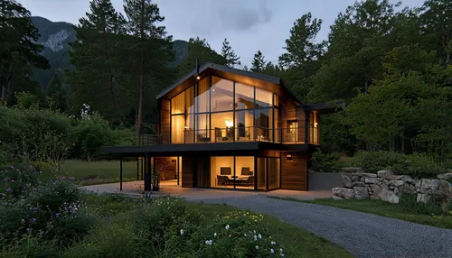 The image shows a house with a modern rustic style, located in a natural setting surrounded by a dense forest. The home features a steeply pitched gable roof and large floor-to-ceiling windows that ma