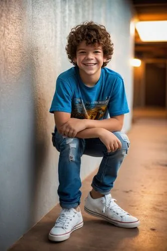 Gaten Matarazzo, young boy, smiling face, bright blue eyes, messy brown hair, casual wear, white graphic t-shirt, light blue jeans, sneakers, relaxed pose, leaning on a wall, indoor setting, warm ligh