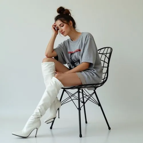 The woman in the image is seated on a black metal chair with a minimalist design. Her posture is relaxed but deliberately arranged for visual effect. She leans slightly to her right, with her right el