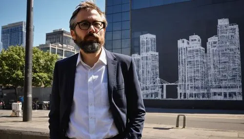 Middle-aged, male, architectural designer, Kettering, standing, hands in pockets, black-framed glasses, short brown hair, beard, white shirt, dark blue jeans, sneakers, holding a roll of blueprints, c