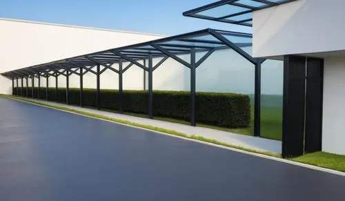 A clean and modern outdoor walkway with a series of metal posts supporting a glass roof. The posts are matte black. The setting features well-manicured hedges on the side, a smooth road with yellow li