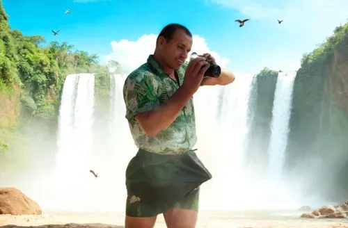 THE MAN IS IN THE AFRICAN JUNGLE. IN THE BACKGROUND THERE IS A WATERFALL WITH SOME SAND, ROCKS AND BIRDS FLYING.
THE MAN IS WEARING GREEN BOXERS. THE MAN'S SHIRT IS OUTSIDE THE BOXERS.,a man taking a 