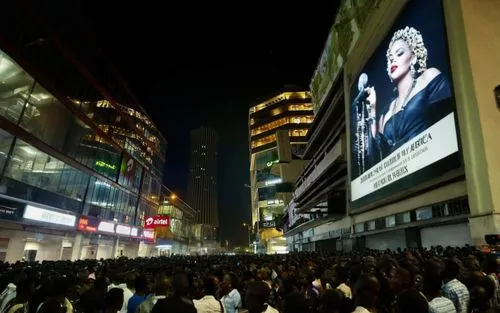 On a building there is a huge sign that says: "MADONNA, WELCOME TO AFRICA". Below a crowd of black boys, men are waiting for Madonna; there are photographers and cameras.,large crowd gathered on the s