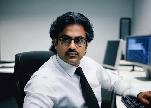 Middle-aged man, Jay Ramachandran, 2002 style hair, glasses, formal wear, suit and tie, sitting in a modern office, desk with multiple monitors, keyboard, and papers, designing security architecture s