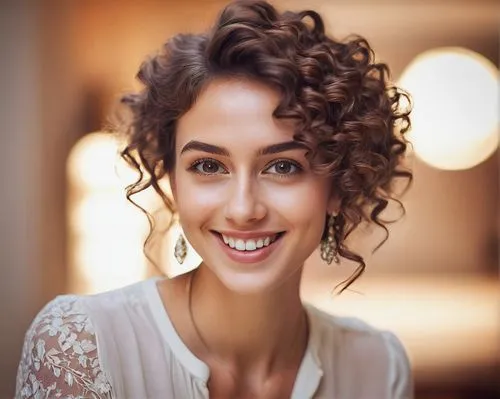 Young adult woman, selfie, smiling, bright eyes, natural makeup, curly brown hair, elegant earrings, white blouse, relaxed shoulders, gentle facial expression, blurred background, soft focus, warm lig