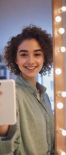 Young woman, selfie-taking, Snapchat filter, bright smile, bold eyebrows, curly brown hair, natural makeup, casual clothes, relaxed posture, phone in hand, mirror reflection, blurred background, soft 