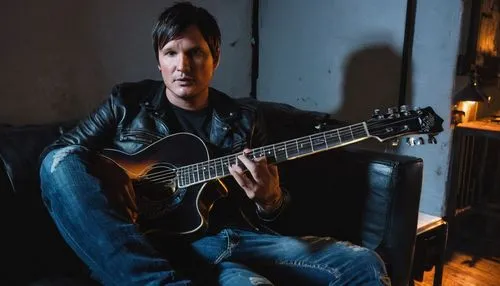 Tom Delonge, Blink-182, musician, solo, (40yo), messy brown hair, few days of beard, black leather jacket, white T-shirt, ripped blue jeans, Converse shoes, holding a guitar, sitting on a couch, backs