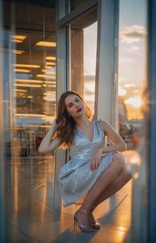 a photography full body of young caucasian woman 26 yers old, brown hair, blue eyes, red lips, whit a hand in the hair wearing a silver dress to knelt in the floor close to a window of a store in a su