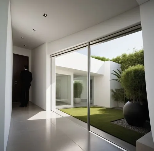 INTERIOR DEL VESTIBULO DE UNA CASA, QUE DA  AUN JARDIN INTERIOR CON MACETAS Y PLANTAS EXOTICAS, CANCALES DE ALUMINIO, CRISTAL, ILUMINACIÓN ELEGANTE Y TENUE,a man stands in the doorway looking out to a