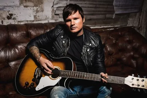 Tom Delonge, Blink-182, musician, solo, (40yo), messy brown hair, few days of beard, black leather jacket, white T-shirt, ripped blue jeans, Converse shoes, holding a guitar, sitting on a couch, backs