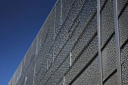 Modern building facade, perforated metal panels, intricate patterns, silver-gray color, industrial aesthetic, urban cityscape, daytime, sunny weather, clear blue sky, few white clouds, sharp angles, g