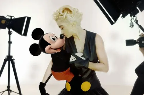 Madonna is on a movie set with Mickey Mouse in her arms!  Madonna is wearing a sexy dress and has beautiful legs. The movie set is full of crew members such as: director, camera operator, sound contro