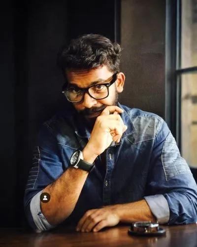 a man wearing a blue shirt and glasses sits at a table with a smart phone,anirudh,kammula,natekar,devarakonda,devvarman,meghnad