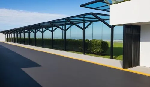 A clean and modern outdoor walkway with a series of metal posts supporting a glass roof. The posts are now matte black. The setting features well-manicured hedges on the side, a smooth road with yello