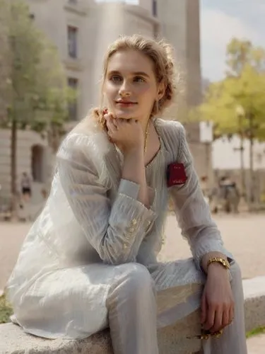 a woman with long curly hair and a golden apron sits on a bench in a park, his eyes fixed on the world beyond. She wears a red dress and a brown t-shirt, and her hair is pulled back into a ponytail. T