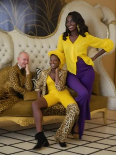 The picture shows three stylishly dressed women posing on an opulent, cream-colored sofa with button-tufting. The background is colorful and vibrant, with plant motifs on the wall adding an exotic tou