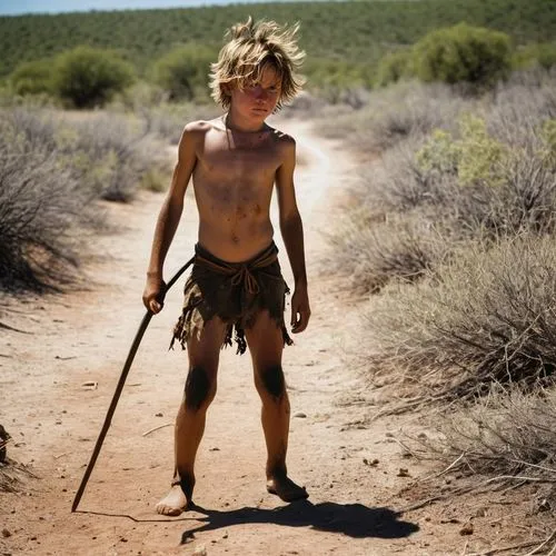 Shirtless sunburn barefoot filthy dirty angry young boy in the wild holding a spear with messy hair covered in sweat dirt and scars in bright sunshine in blazing heatwave sneaking around hunting in th