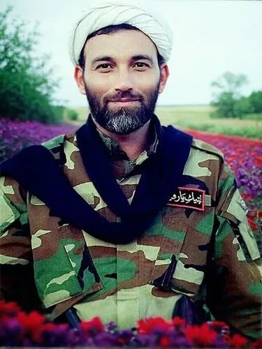 In the plain full of red and purple flowers,the man in camo is wearing a turban and standing near flowers,jalili,soleimani,shahristani,razavi,hayatullah,suleimani,zabiullah,naimatullah,belmokhtar,kurd