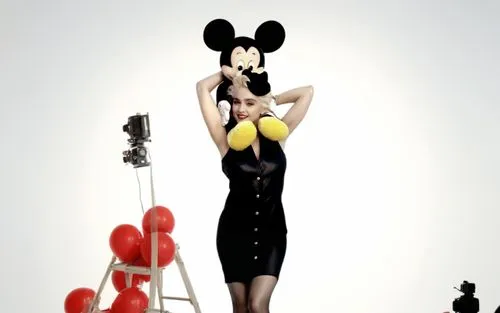 Madonna is on a movie set and she has Mickey Mouse on her shoulders. Madonna is wearing a sexy dress and has beautiful legs and she is wearing high heels. The movie set is full of crew members such as