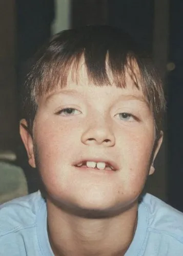 This photo shows the head and shoulders of a young boy. He appears to be around elementary school age, possibly about 10 years old. He has fair skin. His dark brown hair is cut in a straight fringe, c