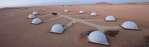 The camp in the desert of KSA contains a group of white geodesic dome tents, and there are lots of landscape lights to define the road between the tents. Every tent has beside it an Arabic seat and a 