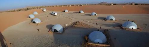 The camp in the desert of KSA contains a group of white geodesic dome tents, and there are lots of landscape lights to define the road between the tents. Every tent has beside it an Arabic seat and a 