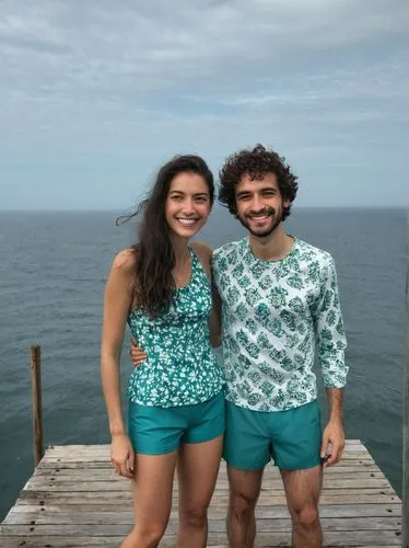 The picture shows two people standing on a wooden jetty by the sea, smiling friendly into the camera.
Both people are wearing the following clothes:
	- The woman is wearing a short dress.
	- The man i