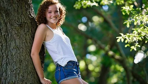 Freckled skin, youthful girl, 18yo, bright smile, natural makeup, curly brown hair, casual wear, white tank top, denim shorts, sneakers, leaning against a tree, hand on hip, relaxed posture, warm sunn